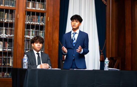 Two young men in suits at a table in a library, one speaking while the other listens.
