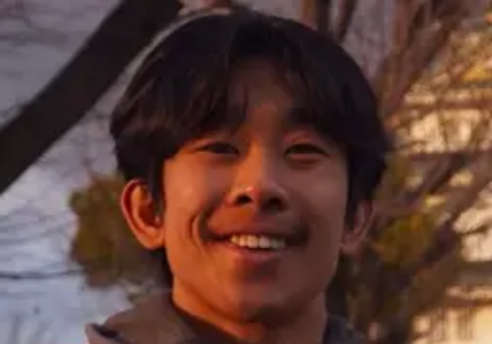Young man smiling outdoors with trees in the background during sunset.