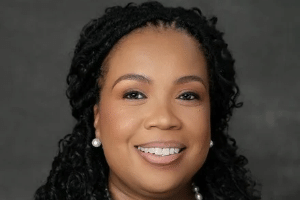 Woman with curly black hair and pearl earrings smiling against a gray background.