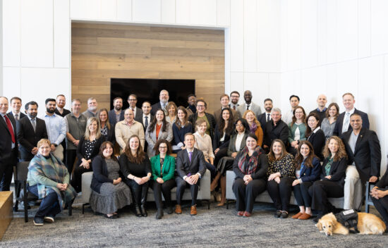 A large group of diverse individuals poses together in a modern interior space, featuring a dog in the foreground.