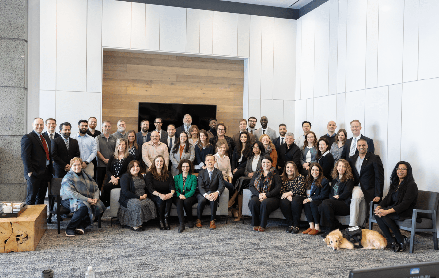 A diverse group of people posing together in an office setting, seated and standing in two rows, with a dog in front.