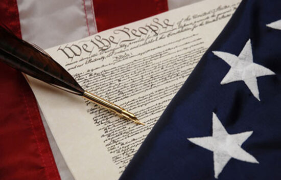 A quill pen resting on the United States Constitution, alongside an American flag.