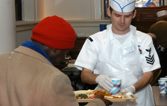 A service member hands a meal and drink to a recipient in a communal dining setting.