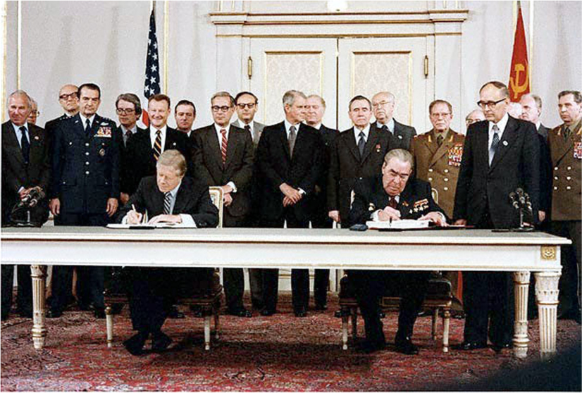 President Jimmy Carter and Soviet leader Leonid Brezhnev sit at a table and sign documents. Officials stand behind them.