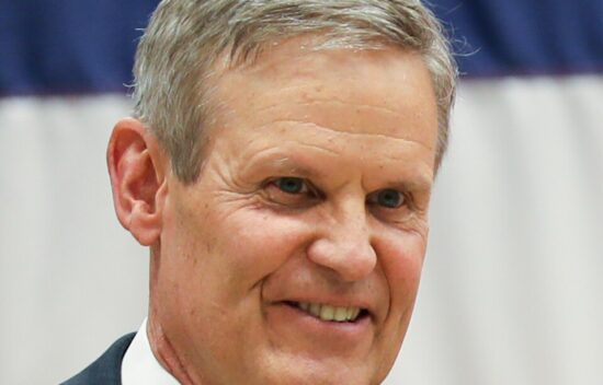 A smiling man in a suit stands in front of a blurred background with a flag.