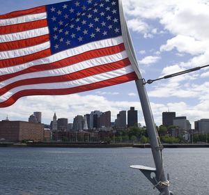 American flag waving in the foreground with a city skyline and cloudy blue sky in the background.