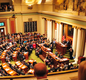 A view of a legislative assembly in session with lawmakers seated and engaging at desks in a grand chamber.