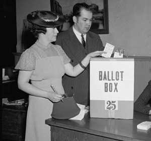 A woman and a man stand at a table, the woman casting a ballot into a box labeled "BALLOT BOX 25."