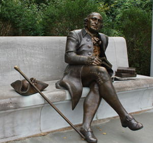 Bronze statue of a seated man in 18th-century clothing, with a cane and a book, on a bench in a park.