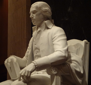 White marble statue of a seated man in historical clothing, holding a book, displayed against a dark background.