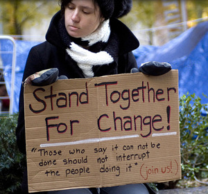 Person holding a cardboard sign that reads "Stand Together For Change!" in an outdoor setting.