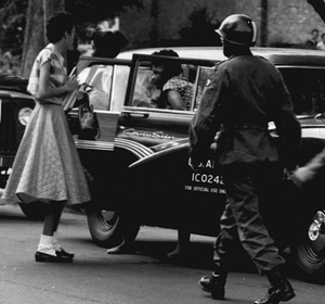 A woman in a skirt stands near a car while a soldier approaches in a historical black and white photo.