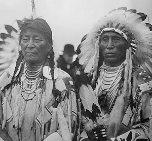 Two Indigenous individuals wearing traditional attire and headdresses, standing closely together against a muted background.
