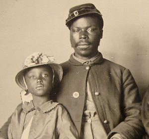 A man and a young girl pose together, both wearing period clothing and hats, in a sepia-toned photograph.