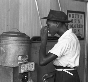 A man drinks from a water fountain labeled "COLORED" in a segregated environment.