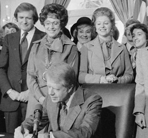A group of people watches as a man signs a document at a formal event.