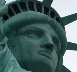 Close-up of the Statue of Liberty’s face, featuring her crown and serious expression, against a light sky.