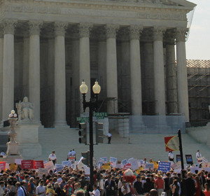 Crowd of protesters with signs gathered in front of a large building with columns under construction.