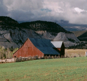 Red barn in a grassy field, surrounded by mountains and a cloudy sky.