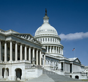 U.S. Capitol building with a dome and columns, under a blue sky with a flag visible on the right.
