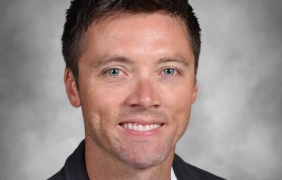 Headshot of a smiling man with short dark hair, wearing a black blazer over a white shirt. Neutral gray background.