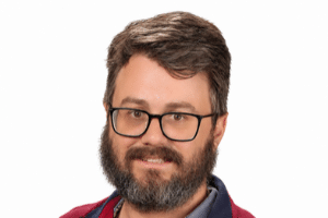A man with glasses and a beard smiles for the camera against a plain background.