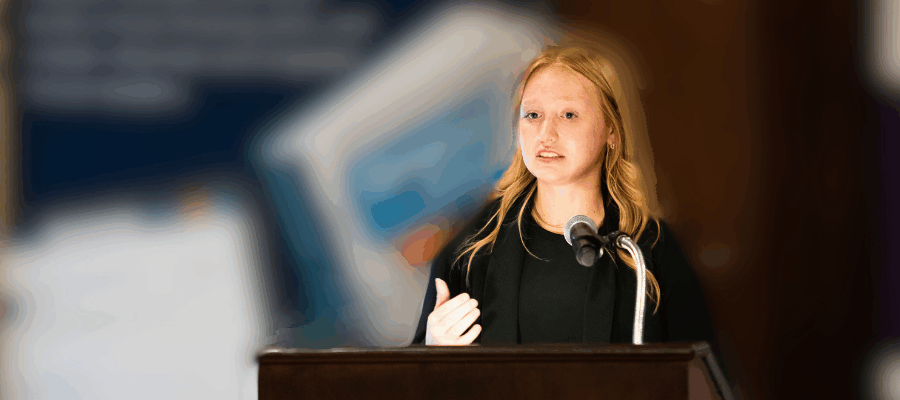 A young woman speaks at a podium with a microphone in a blurred background setting.