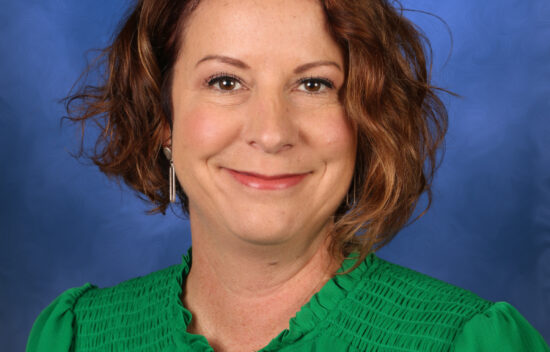 A woman with curly brown hair wearing a green shirt, smiling against a blue background.