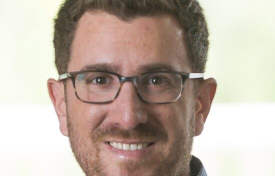 A man with short, curly hair wearing glasses and a blue checkered shirt, smiling at the camera.