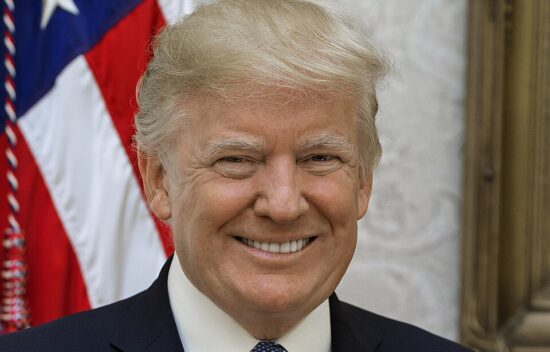 Portrait of a man with light hair, smiling, standing in front of a flag.