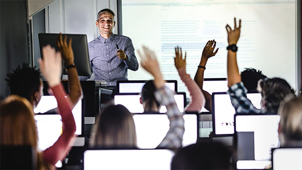 A teacher stands in front of a class with students raising their hands, seated at computer desks.