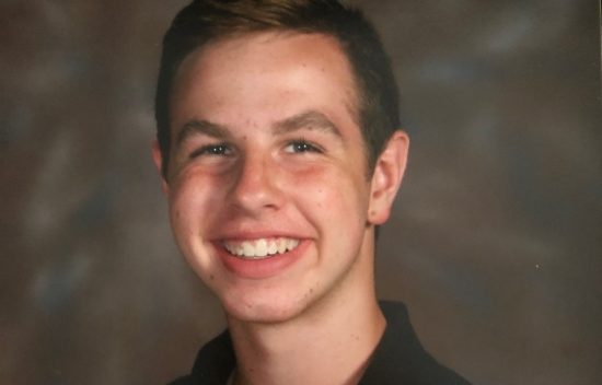 A young man with short brown hair smiles in front of a blurred brown background. He is wearing a black shirt.