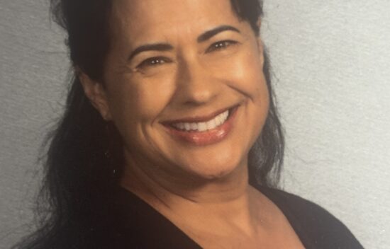 A smiling woman with long dark hair, wearing a black top, against a gray background.