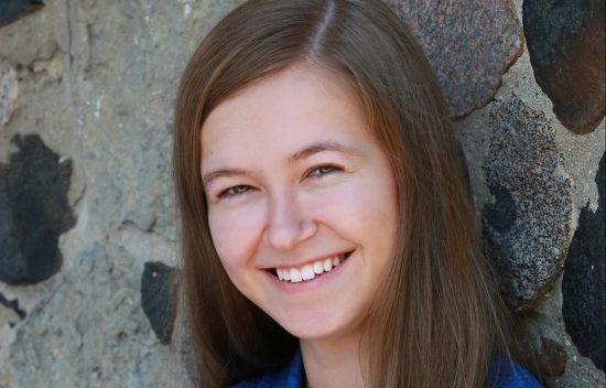 Young woman with long brown hair smiling against a stone wall background. She is wearing a blue top.