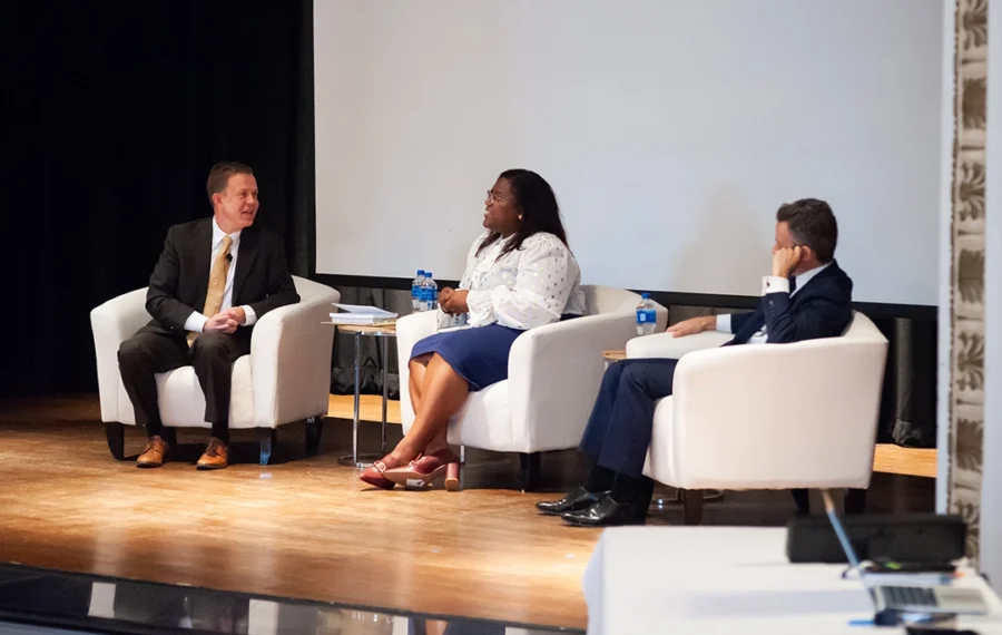Three speakers seated on stage during a panel discussion in a well-lit conference room.