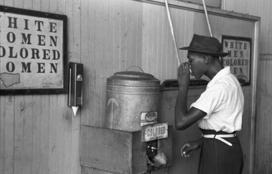 A young Black man drinking from a water fountain labeled "colored" in a racially segregated setting.