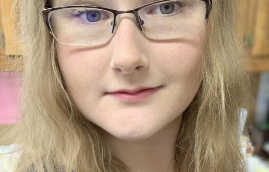 A close-up portrait of a person with long, light hair and glasses, smiling softly at the camera.