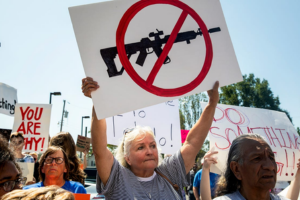 A woman holds a sign with a crossed-out rifle symbol at a protest, surrounded by other demonstrators.
