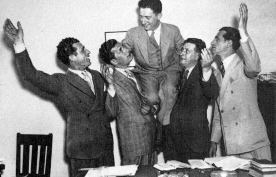 Five men in suits celebrating indoors, with one man being lifted by the others, expressing joy.