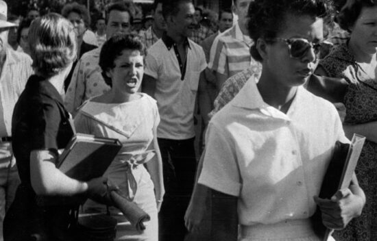 Black and white photo of a diverse group of people, with a focus on a woman holding books, amid a tense crowd.