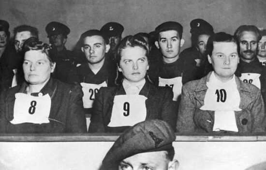 Black and white photo of a group of individuals sitting in rows, some wearing numbers on their shirts, with soldiers in the background.