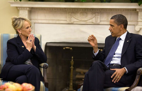 Two individuals engaged in conversation in a formal setting with a fireplace in the background.