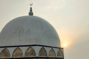 Dome of a mosque with a crescent moon finial, set against a hazy sunset sky.