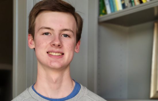 Young man smiling in a casual gray sweatshirt, standing next to a bookshelf with visible books.