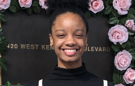 Young woman smiling in front of a floral backdrop with pink roses and a sign reading "420 WEST KENTUCKY BOULEVARD."