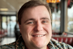 A smiling person in a casual setting with soft lighting and restaurant decor in the background.