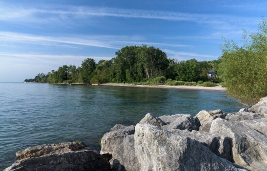Rocky shoreline with calm water, sandy beach, and green trees under a blue sky.