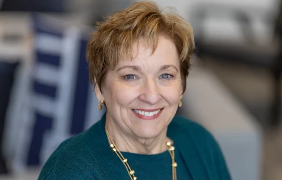 Smiling woman with short, light brown hair, wearing a teal sweater and necklace, in a soft-focus background.