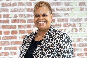 Smiling woman with short hair wearing a leopard-print blazer, standing in front of a brick wall.