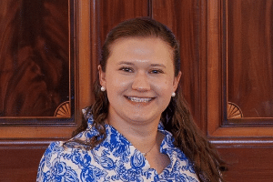 A woman with long brown hair smiles, wearing a blue patterned blouse, posed in front of a wooden background.